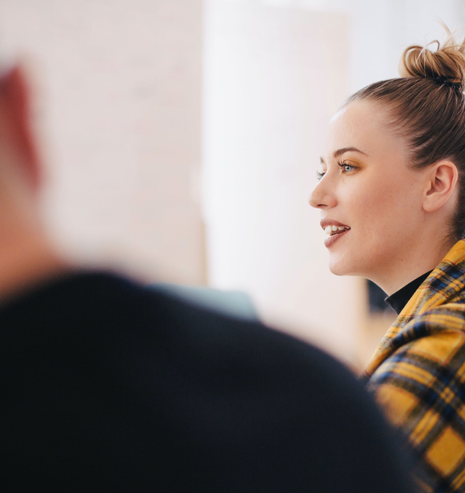 Clear photogaph of woman's side profile speaking with her hair in a bun and wearing a yellow plaid shirt. A man featured slightly out of frame - Usercentrics